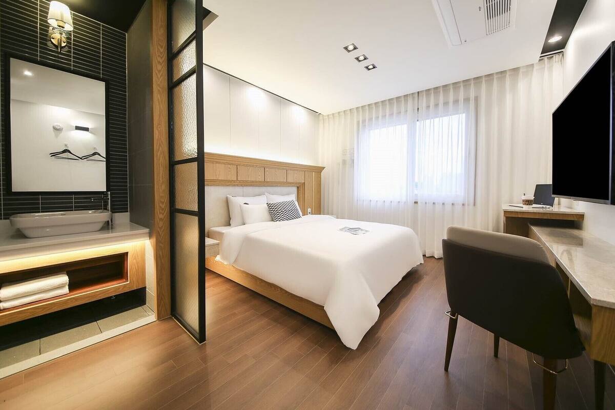 A modern hotel room with a large bed, a desk with a laptop, a TV, and a bathroom with a sink and mirror.