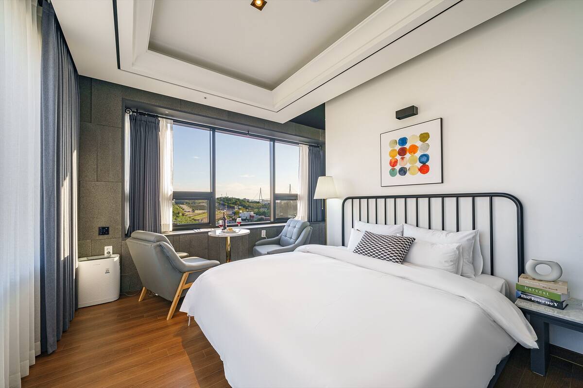 A hotel room with a large bed, a desk with a chair, a window with curtains, and a view of the city.