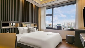 A modern hotel room with a large bed, a desk with a laptop, a TV, and a view of a cityscape. - Urban (Mokpo)