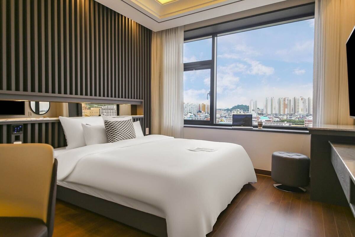 A modern hotel room with a large bed, a desk with a laptop, a TV, and a view of a cityscape.