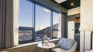 A modern hotel room with a large window offering a city view, a comfortable seating area, a small table with a bottle and glasses, and a lamp. - Urban (Mokpo)