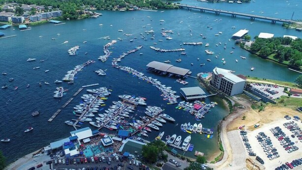 An aerial view of a marina with numerous boats docked, a large building with a flat roof, and a parking lot with cars. - Truly the Best Location & Sunsets on the Lake! Dining & Shopping 2 minutes away! (Osage Beach)