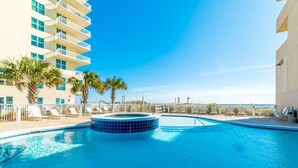 Una piscina con hidromasaje, rodeada de sillas de descanso y palmeras, con un edificio al fondo.