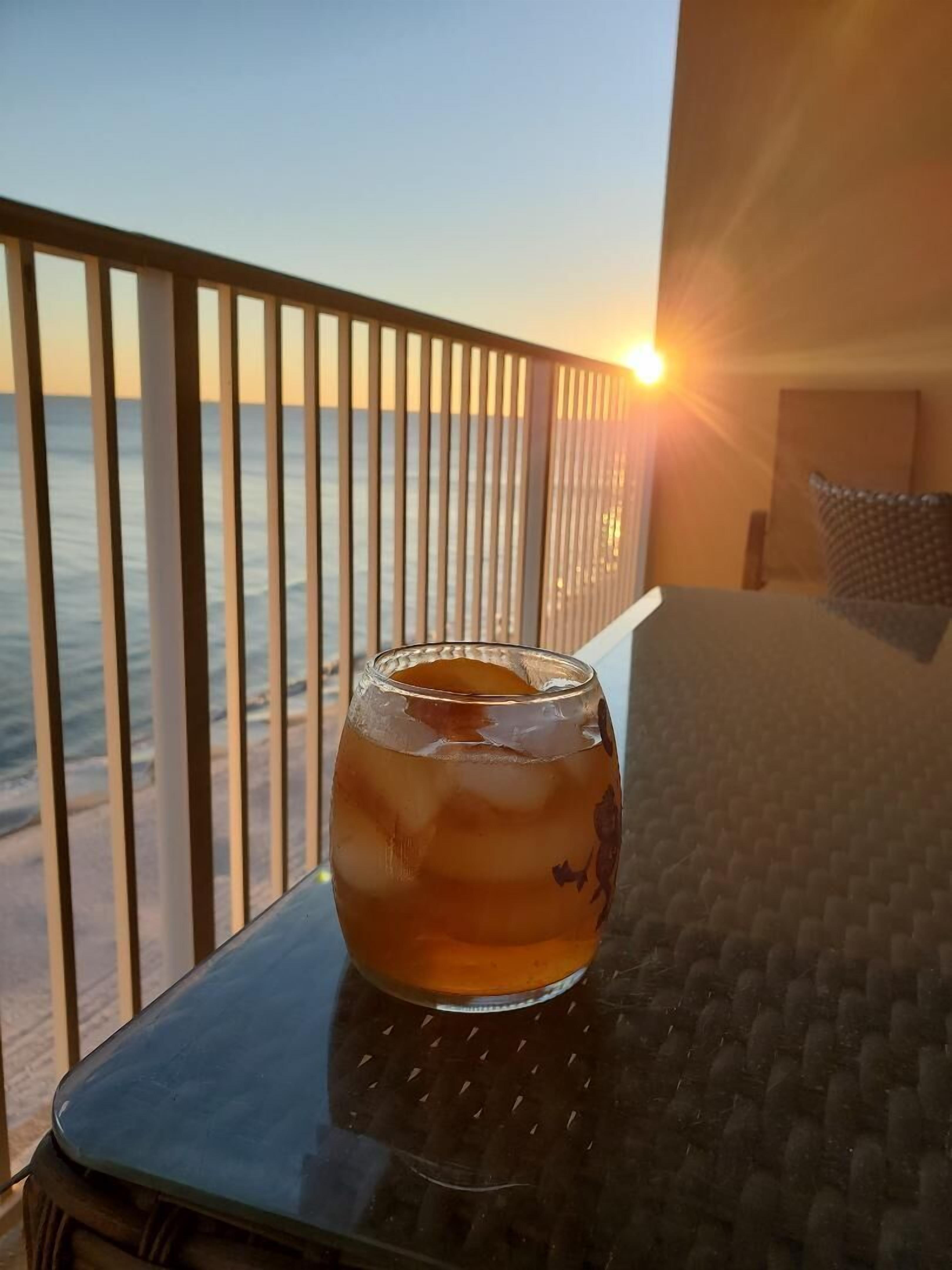 Ein Glas bernsteinfarbene Flüssigkeit auf einem Tisch neben einem Balkon mit Blick auf das Meer bei Sonnenuntergang.