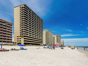 A beachfront hotel with multiple floors, a sandy beach in the foreground, and a clear blue sky.