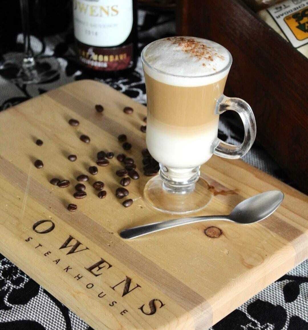 A glass of layered beverage on a wooden board with coffee beans and a spoon. The board has "OWENS STEAKHOUSE" written on it.