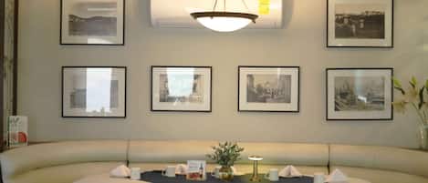 Un restaurante con una larga banqueta curva, cuadros en las paredes y una mesa central con un jarrón pequeño y una lámpara.