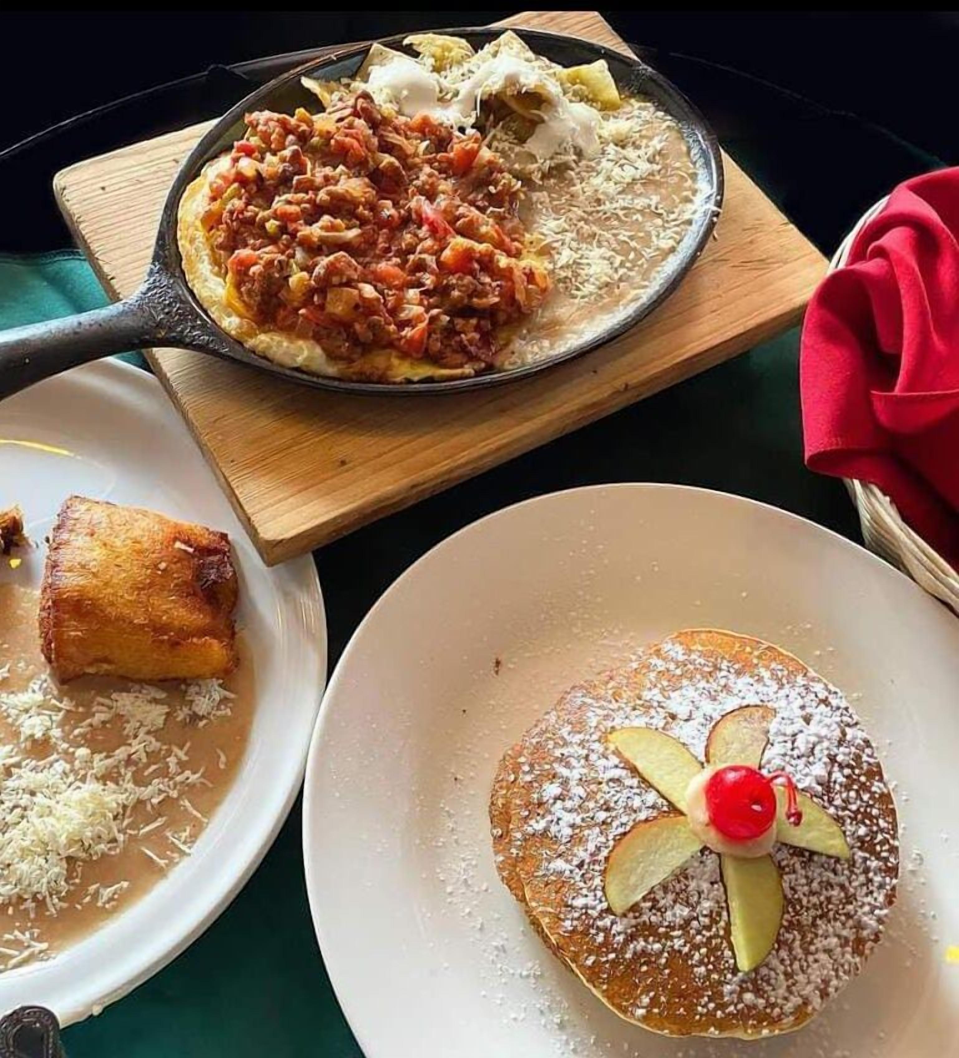 A dining table with a variety of dishes including a skillet with a tomato-based dish, a plate with a dessert topped with a cherry and apple slices, and a plate with a piece of bread.