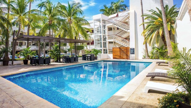 A swimming pool with lounge chairs and a covered seating area, surrounded by palm trees and a multi-story building. - Wyndham Garden Los Mochis Plaza Inn (Los Mochis)