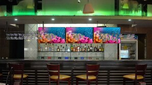 A bar area with multiple seating arrangements, a variety of bottles on the shelves, and a large screen displaying colorful visuals. - Wyndham Garden Los Mochis Plaza Inn (Los Mochis)