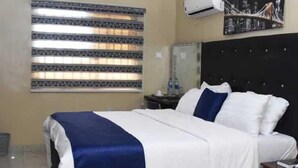 Una habitación de hotel con cama, mesita de noche, ventana con persianas, equipo de aire acondicionado y un cuadro con una vista urbana.