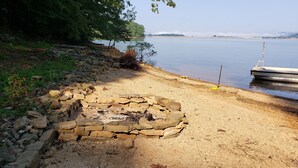 A stone fire pit surrounded by a gravel area near a calm lake with a dock and trees in the background.