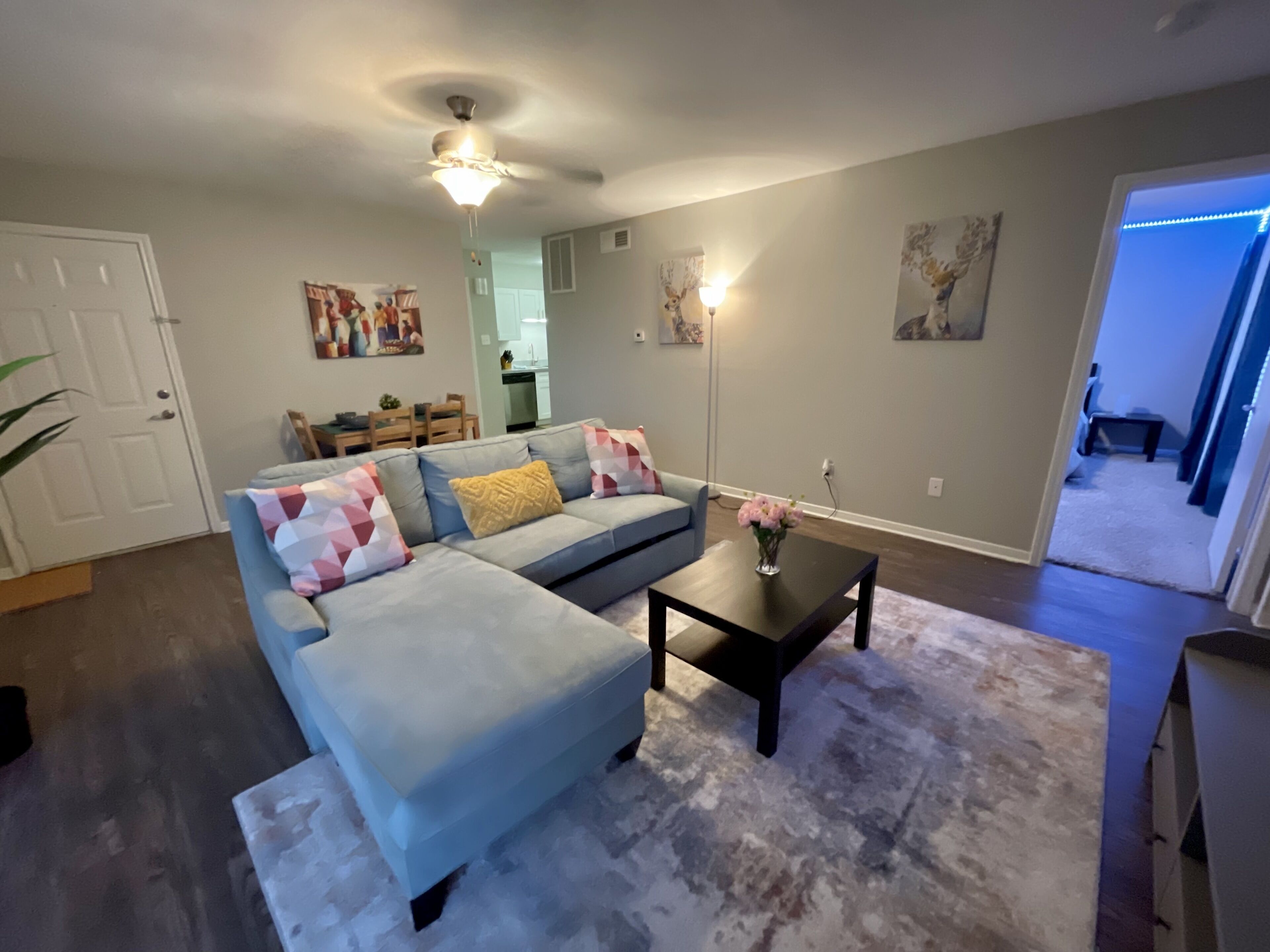 A living room with a light blue sectional sofa, a dark coffee table, and a white rug. There is a ceiling fan, a wall-mounted light, and a door leading to another room.