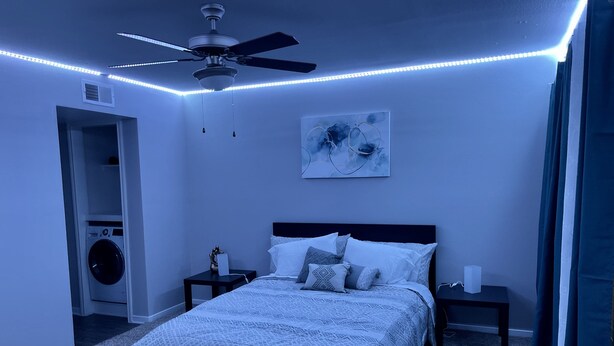 Quarto com cama, mesas de cabeceira, ventoinha no teto, obra de arte na parede e uma máquina de lavar no canto.