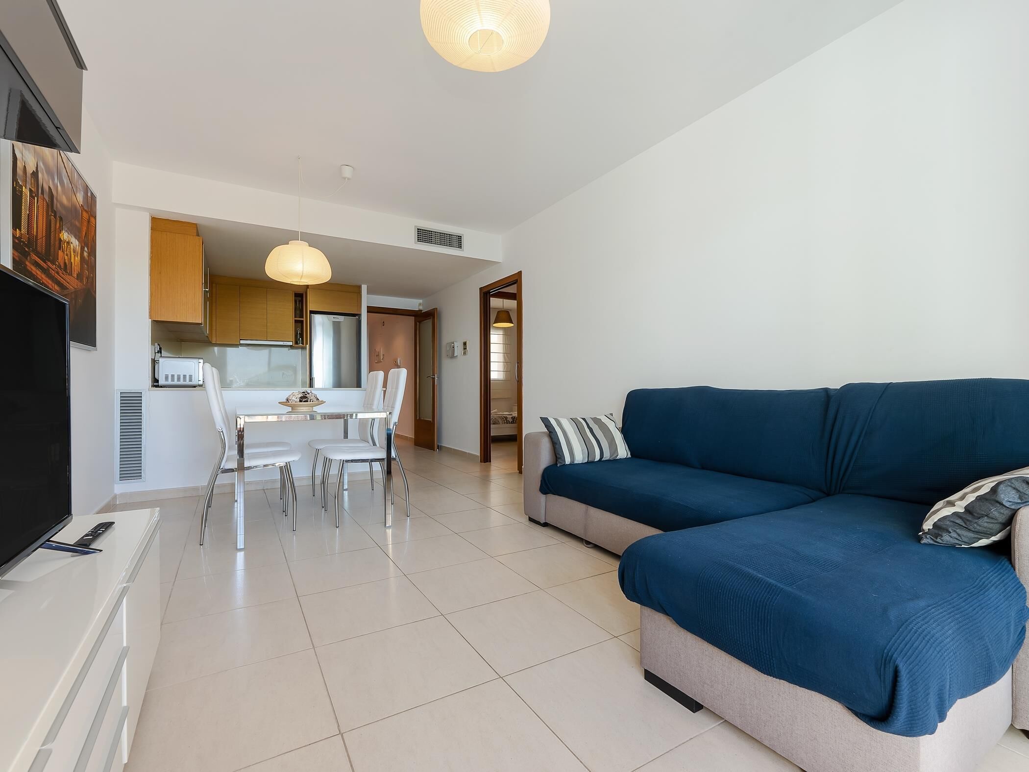 Un saló modern amb un sofà blau, una zona de menjador amb taula i cadires blanques, i una cuina amb armaris de fusta.