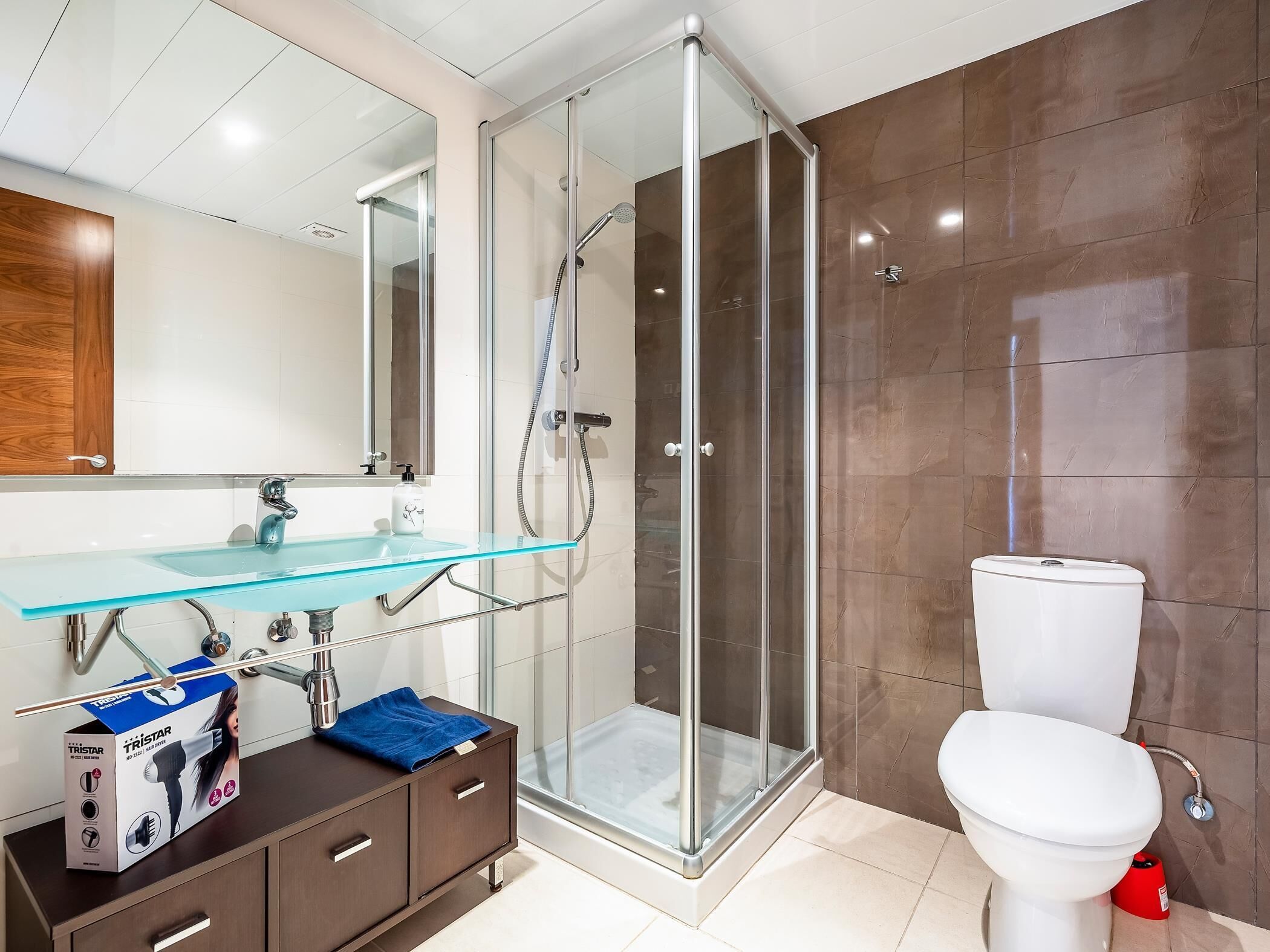 Une salle de bain moderne avec une douche à parois de verre, des toilettes blanches et un lavabo en verre, surmonté d’un meuble en bois.