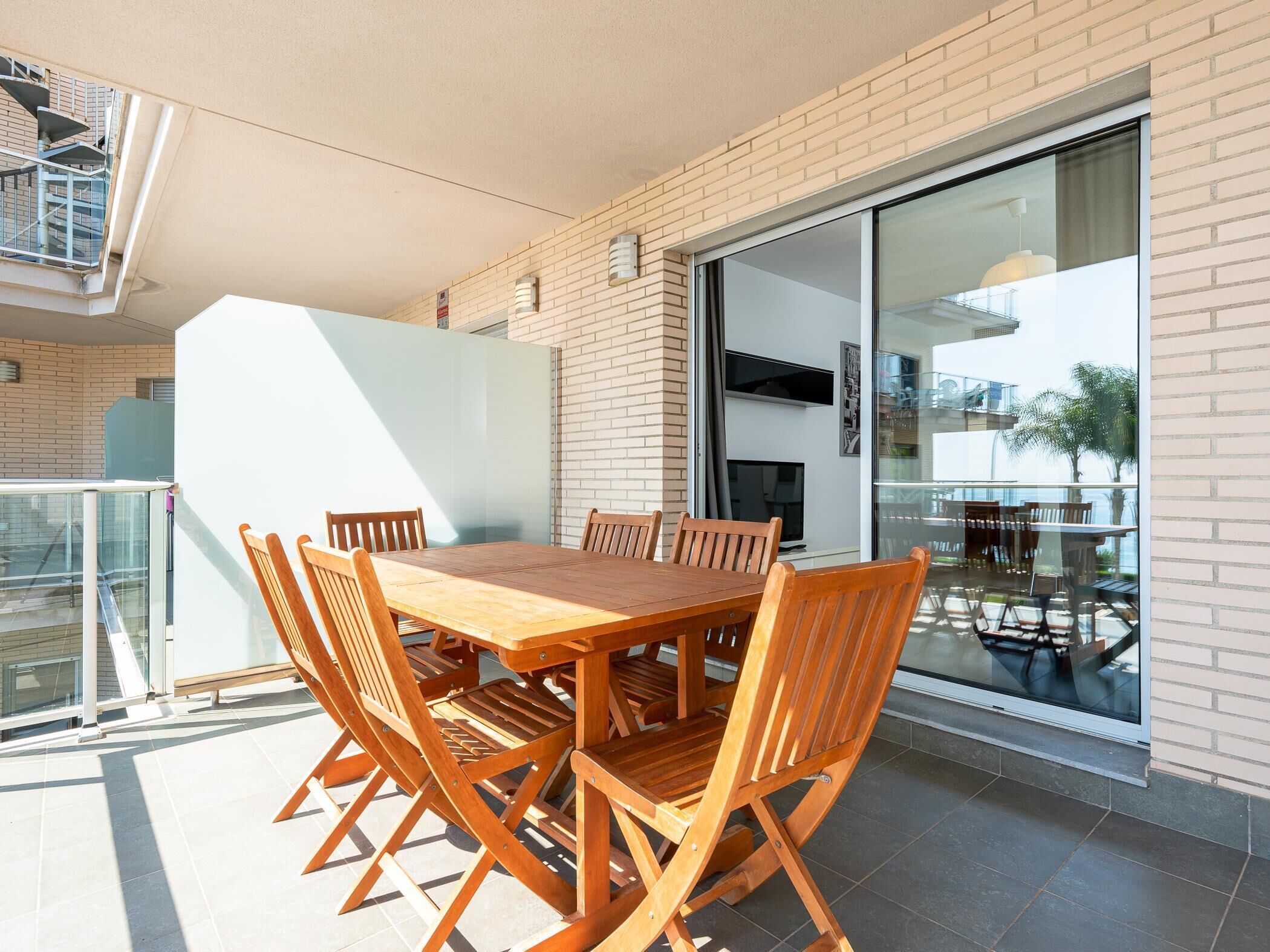 Een houten eettafel met zes stoelen op een balkon, met een glazen deur, een televisie en uitzicht op een gebouw met balkon.