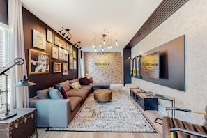 Uma sala de estar moderna com um sofá marrom, um banquinho redondo, um tapete estampado e uma parede de tijolos com o letreiro 'Hard Rock Cafe'.