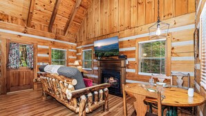 A log cabin interior with a wooden sofa, a fireplace, a dining table, and a TV mounted above it.