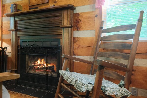 A rustic cabin interior with a fireplace, wooden walls, and a wooden chair with a checkered cloth.
