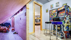 Een kamer met een schuine balken plafond, houten wanden en een zichtbare deur naar een andere ruimte. Er staan twee metalen krukken met een blauwe zitting, een klein tafeltje en verschillende speelgoed op de vloer.