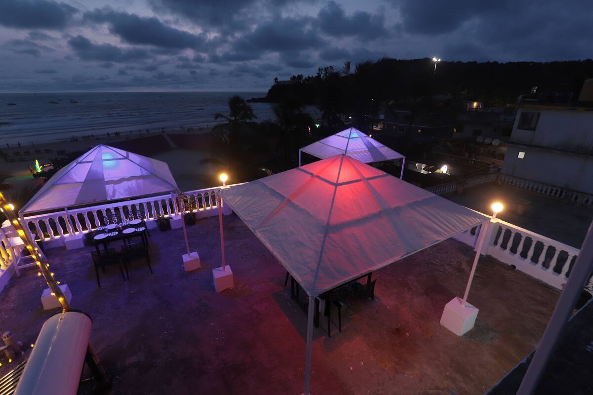 A rooftop terrace with two large, transparent canopies providing shade. The terrace is equipped with tables and chairs, and there are string lights along the railing. The background features a view of the ocean and a distant shoreline.