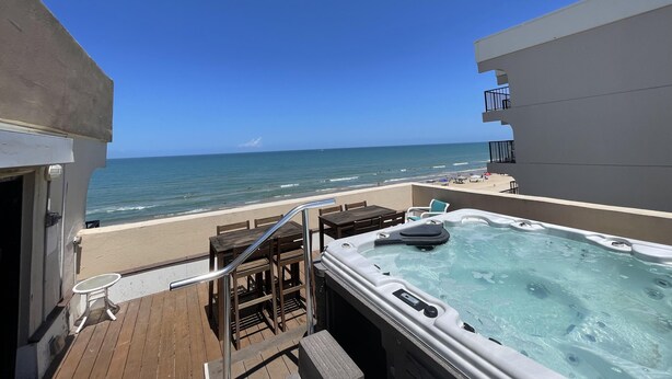 A deck with a hot tub, wooden furniture, and a view of the ocean. - Beachfront Penthouse, private rooftop balcony & jacuzzi (South Padre Island)