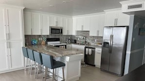 A modern kitchen with white cabinetry, a marble countertop, stainless steel appliances, and bar stools. - Beachfront Penthouse, private rooftop balcony & jacuzzi (South Padre Island)