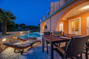 Uma área à beira da piscina com espreguiçadeiras, uma mesa com bebidas e uma parede de pedra.