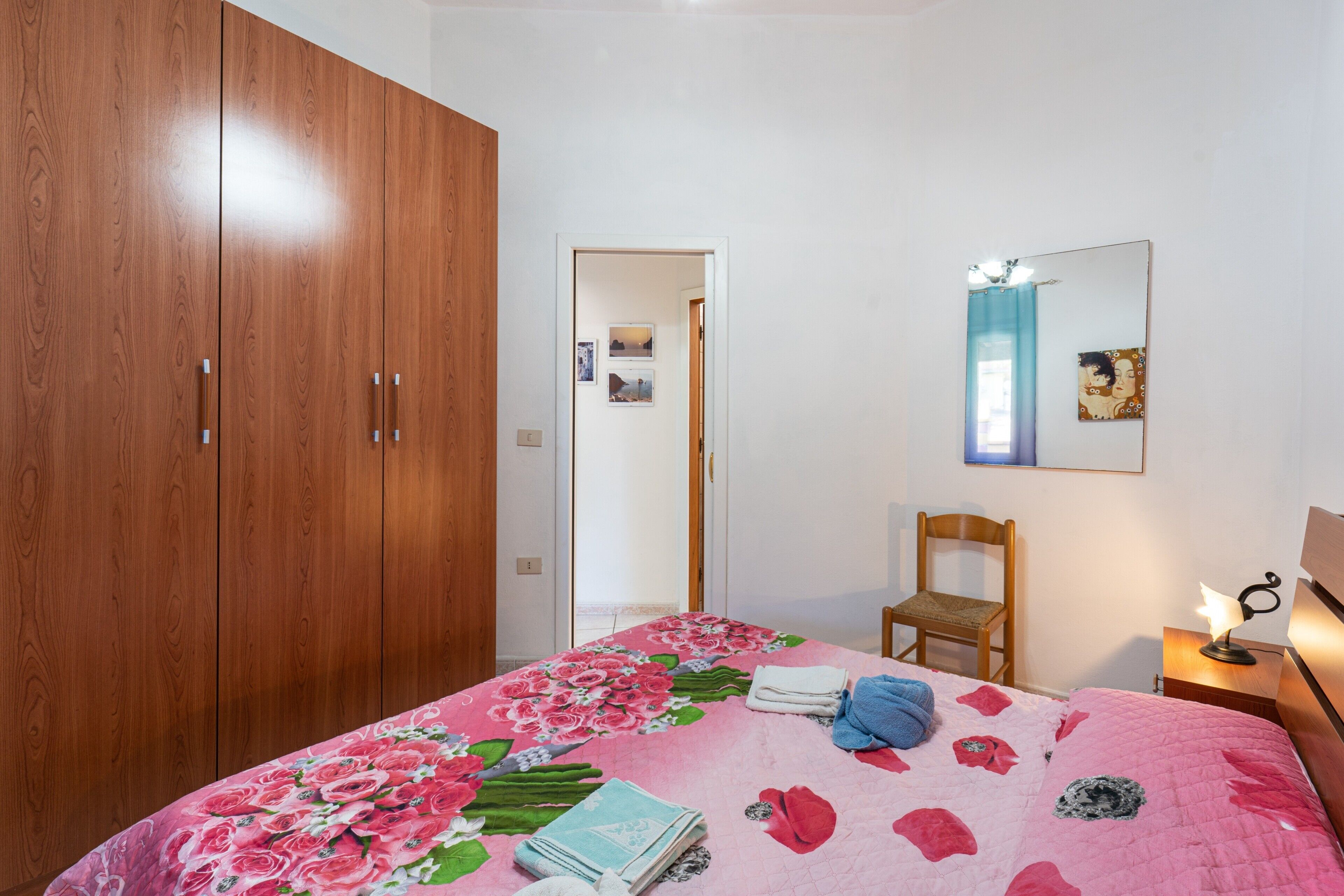 Una camera da letto con un'armadiatura in legno, un letto con copripiumino floreale, una sedia, un comodino con una lampada e uno specchio a parete.