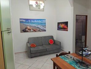 A room with a grey sofa, a dining table with a blue table runner, and two wall-mounted pictures depicting a beach and a colorful splash.