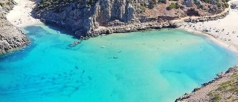 A coastal landscape with a clear turquoise bay, rocky terrain, and sparse vegetation.