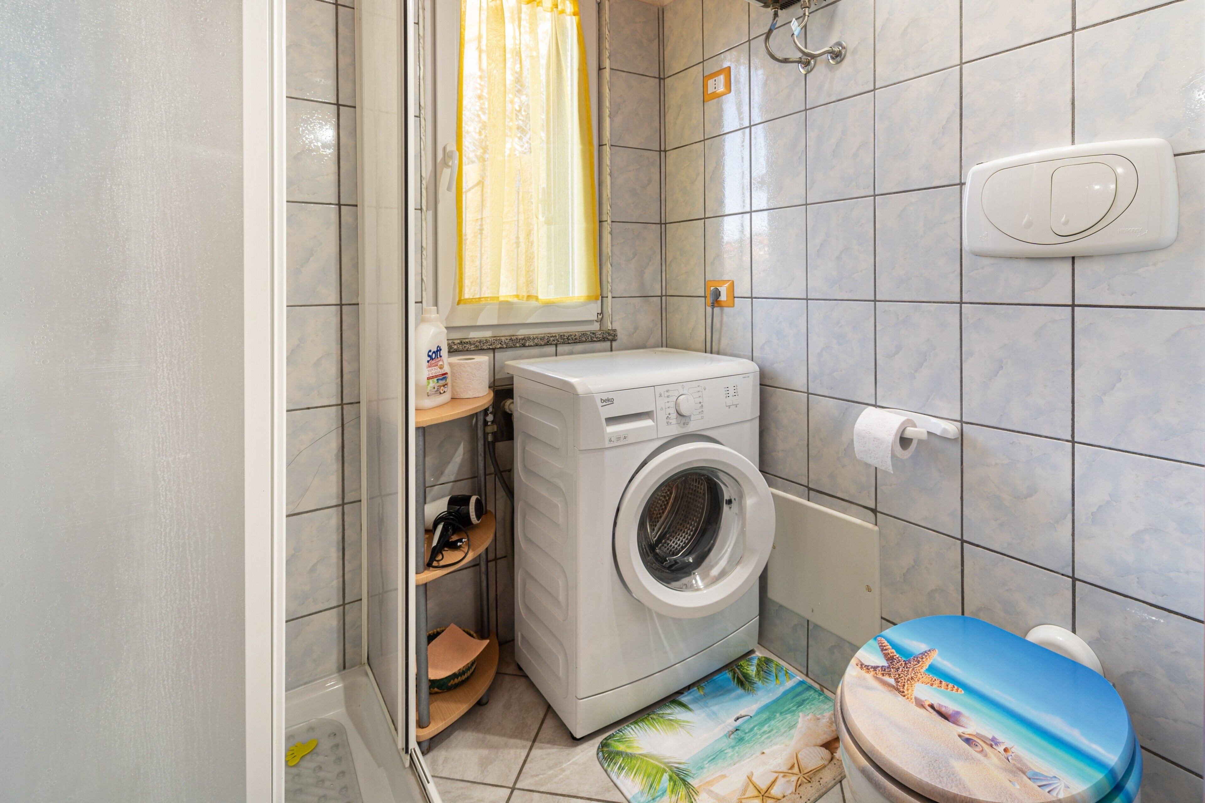Un bagno compatto con lavatrice, WC con coperchio a tema spiaggia e finestra con tenda.