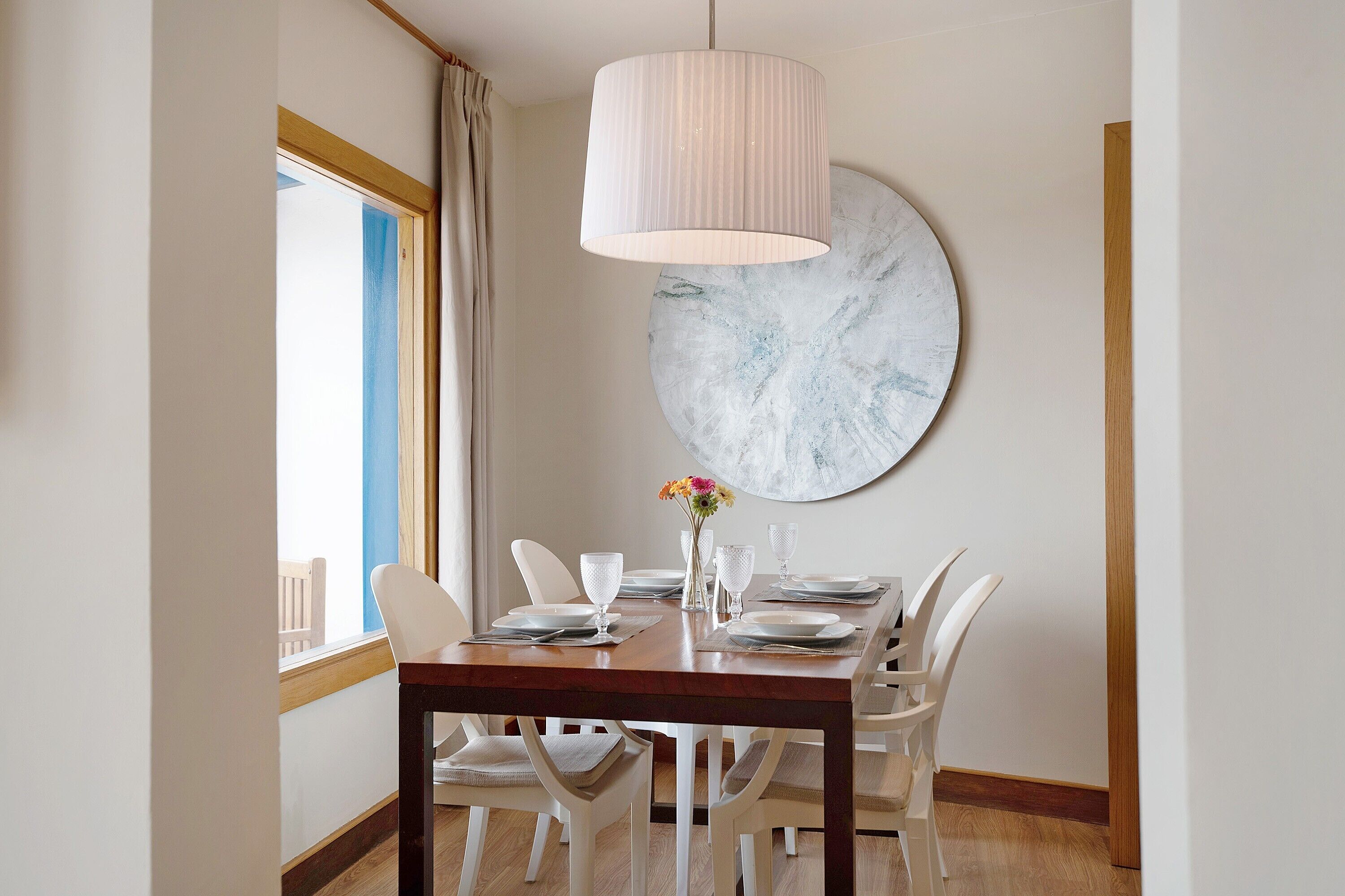 Een eetruimte met een houten tafel, witte stoelen, een hanglamp en een groot abstract schilderij aan de muur.
