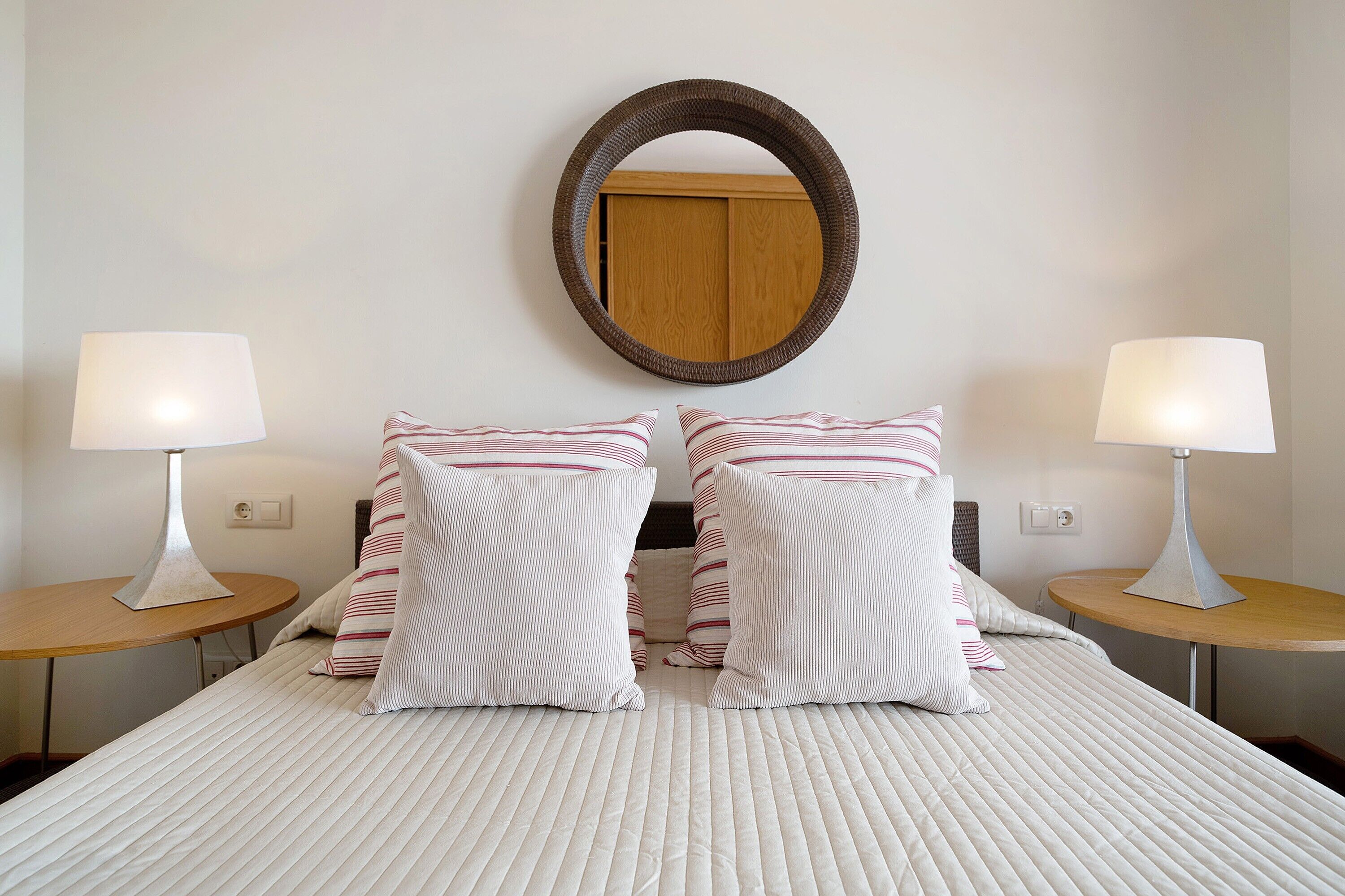 A neatly made bed with striped pillows, two bedside tables with lamps, and a wooden wardrobe in the background.