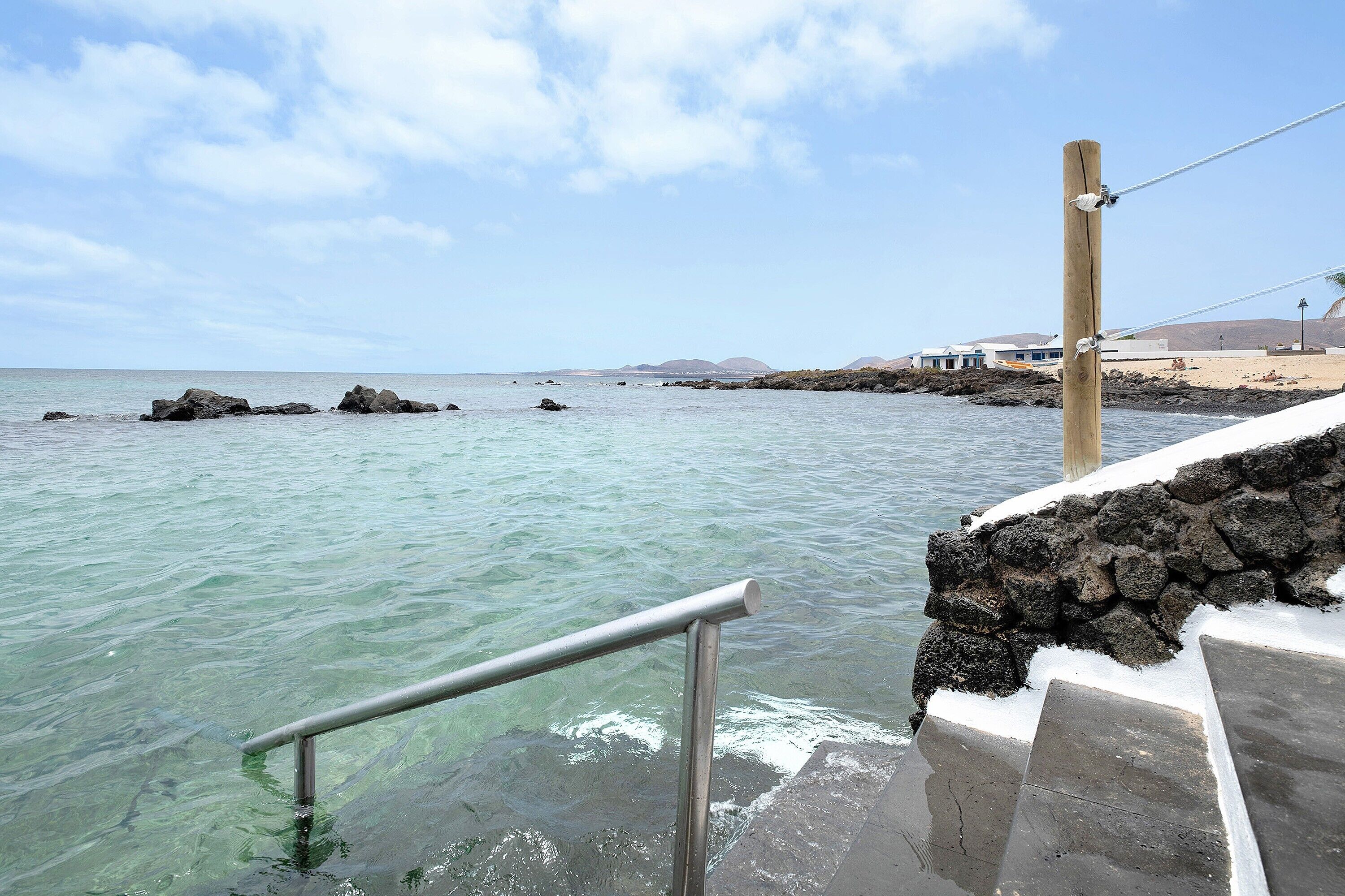 A coastal walkway with a metal railing, steps leading down to the water, and a rocky shoreline in the background.