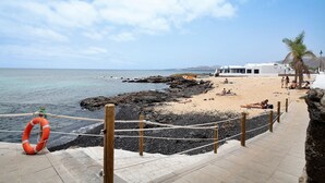 Un paseo marítimo costero con un aro salvavidas, costa rocosa, playa y un edificio de fachada blanca.