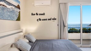 Un dormitorio con cama, almohadas, aire acondicionado de pared y vista al mar desde un balcón.