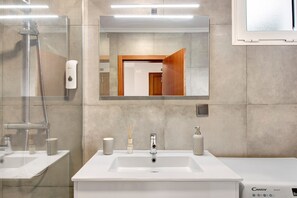 Un baño moderno con ducha de vidrio, un lavamanos blanco con dispensador de jabón y un espejo sobre el lavamanos.