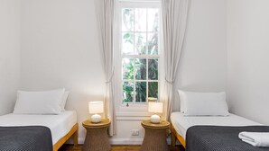Een kamer met twee eenpersoonsbedden, elk met een wit kussen en een donkere deken. Twee rieten nachtkastjes met lampen staan ernaast, en er is een raam met gordijnen.