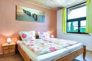 Una cama doble con ropa de cama floral, una mesita de noche con lámpara, un cuadro en la pared y una ventana con cortinas.