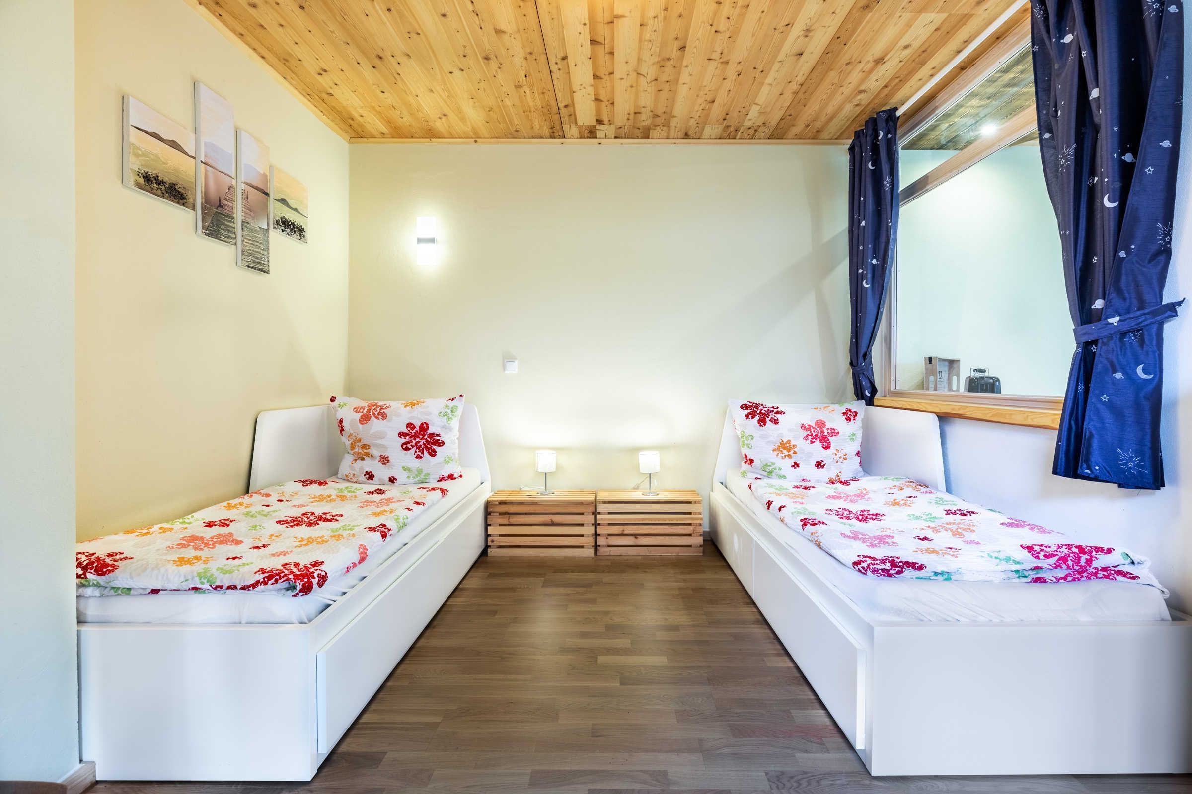 Une chambre avec deux lits, un plancher en bois, un plafond en bois et une fenêtre avec des rideaux.