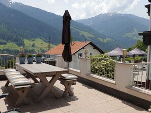 A terrace with a table and benches, potted plants, and a view of mountains.