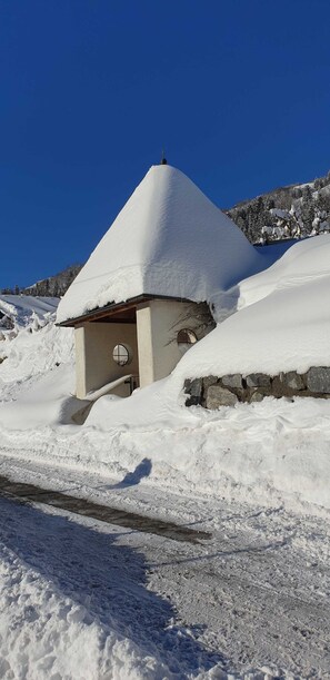 A snow-covered building with a conical roof, a circular window, and a stone wall.