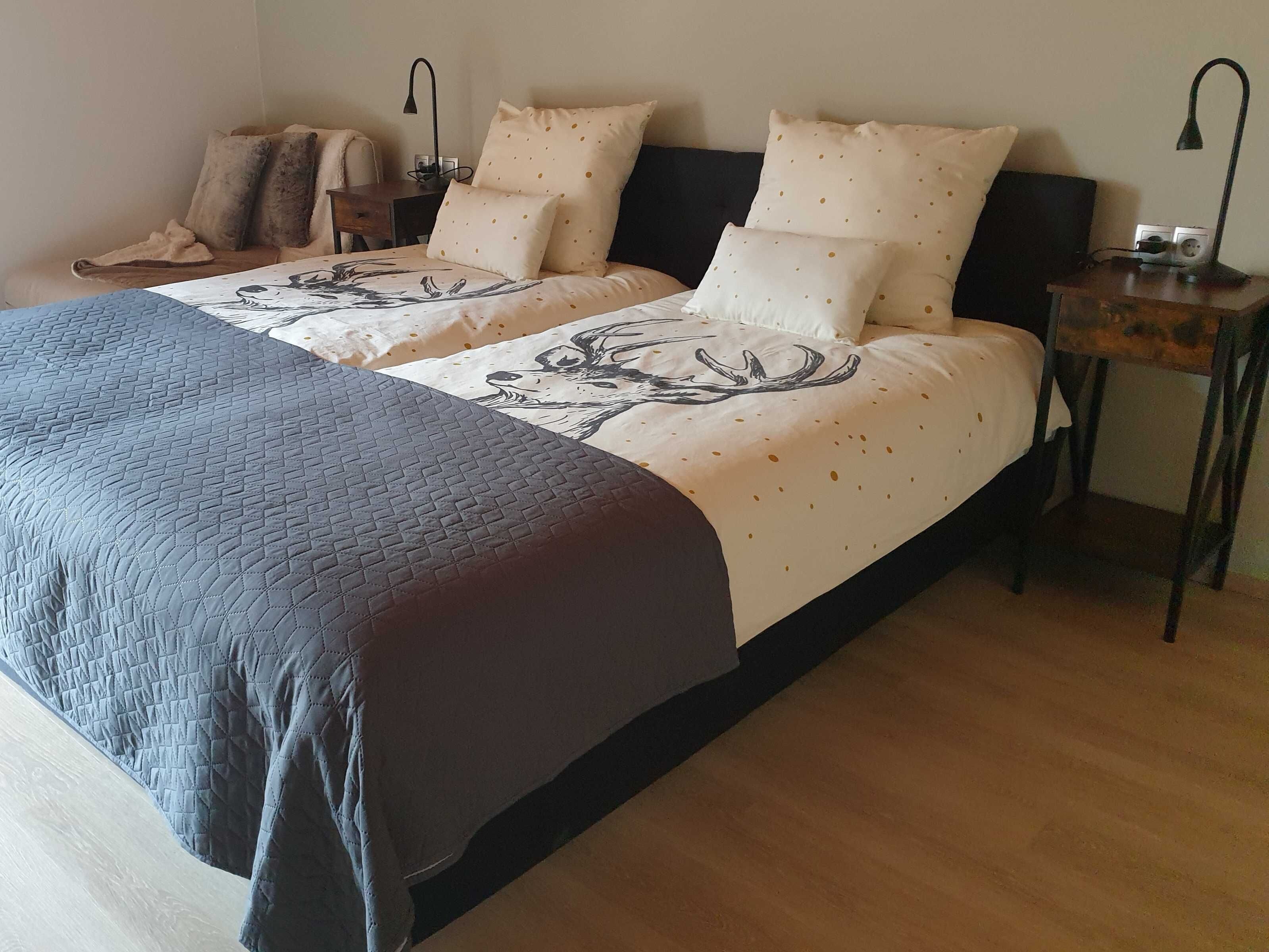 A double bed with a quilted cover, two bedside tables with lamps, and a chair with a blanket.