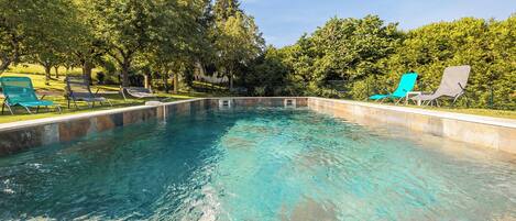 A rectangular swimming pool with clear blue water, surrounded by a stone border. There are lounge chairs along the pool's edge, and trees in the background.