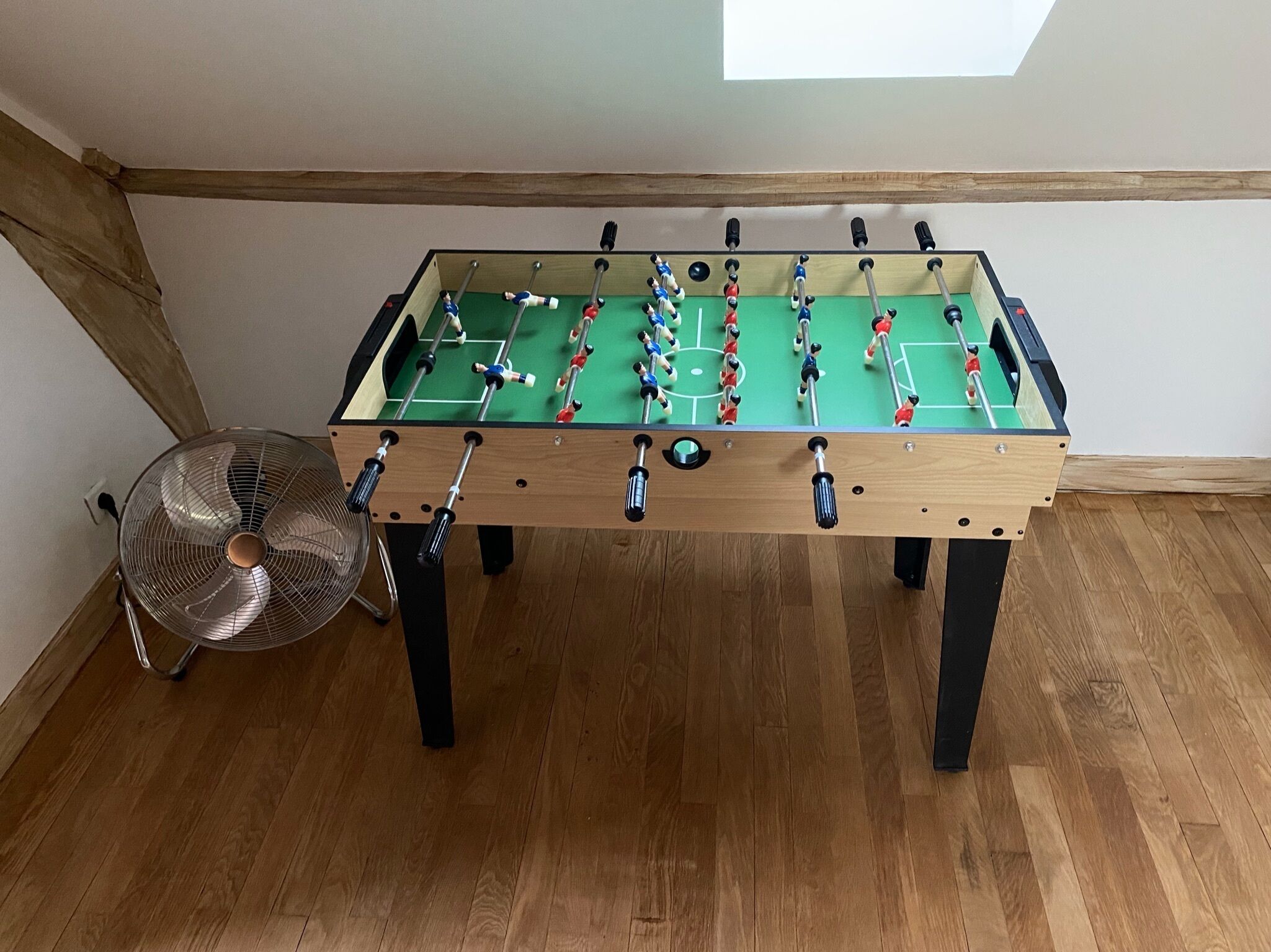 Une table de baby-foot avec un ventilateur à proximité, un plancher en bois et un plafond en pente.