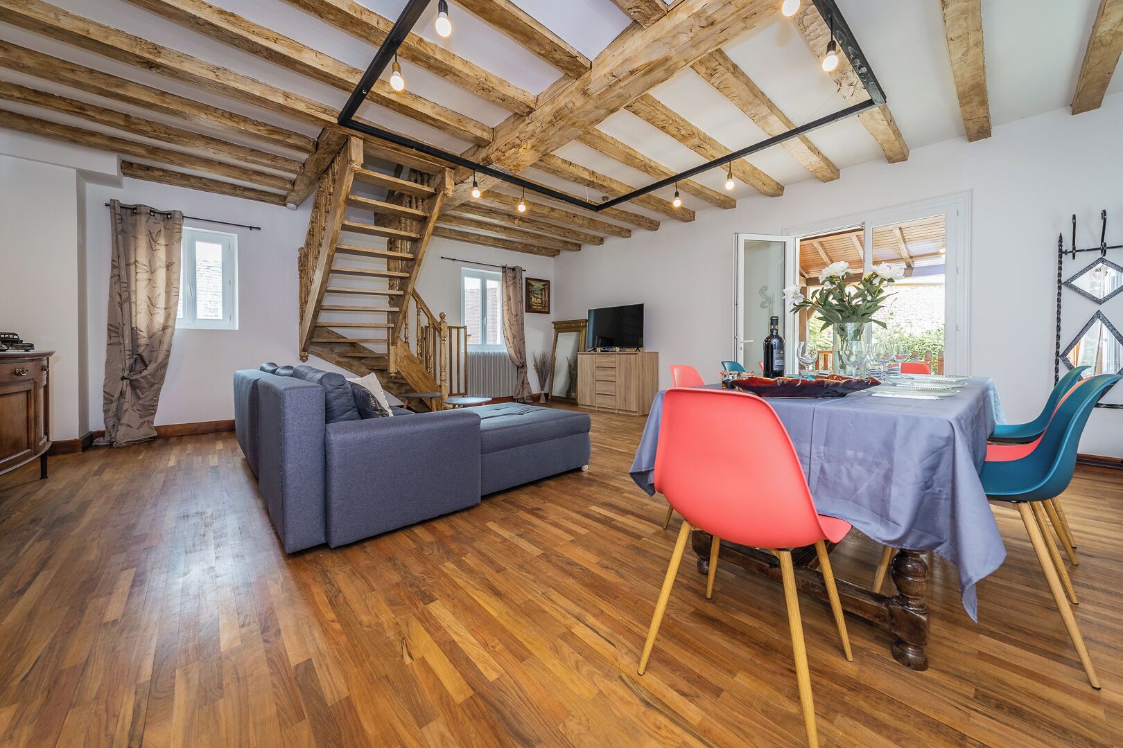 Un vaste espace de vie doté d’un plafond en bois, d’un canapé gris, d’une table à manger avec des chaises et d’un escalier.