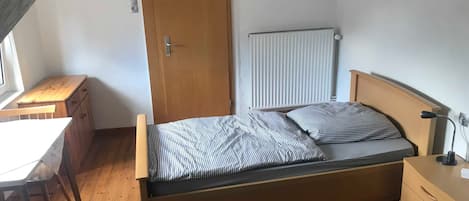 A bedroom with a wooden bed, a radiator, a wooden dresser, a desk, and a window.