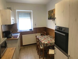 Eine kompakte Küche mit Esstisch, einer Kaffeemaschine, einem Backofen und einem Fenster mit Jalousien.